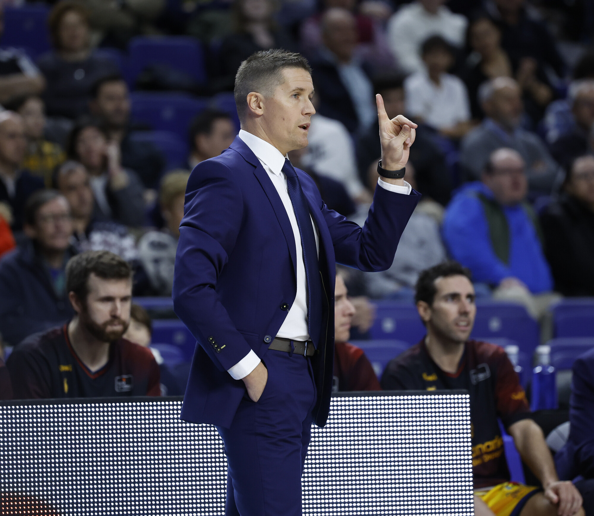 Entrenador gesticulando durante un partido, con público en el fondo.