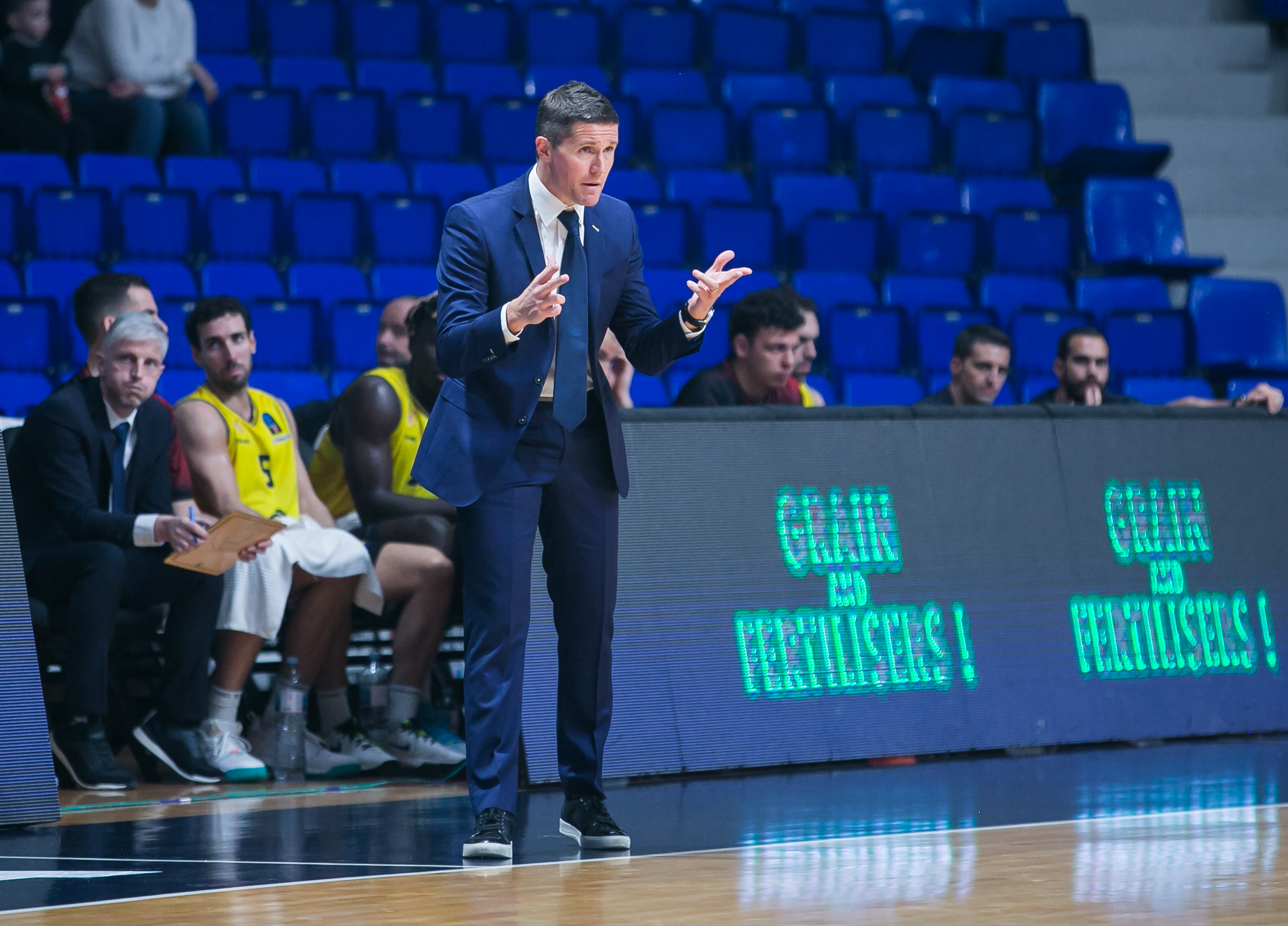Entrenador gesticulando durante un partido, con jugadores en el banquillo.