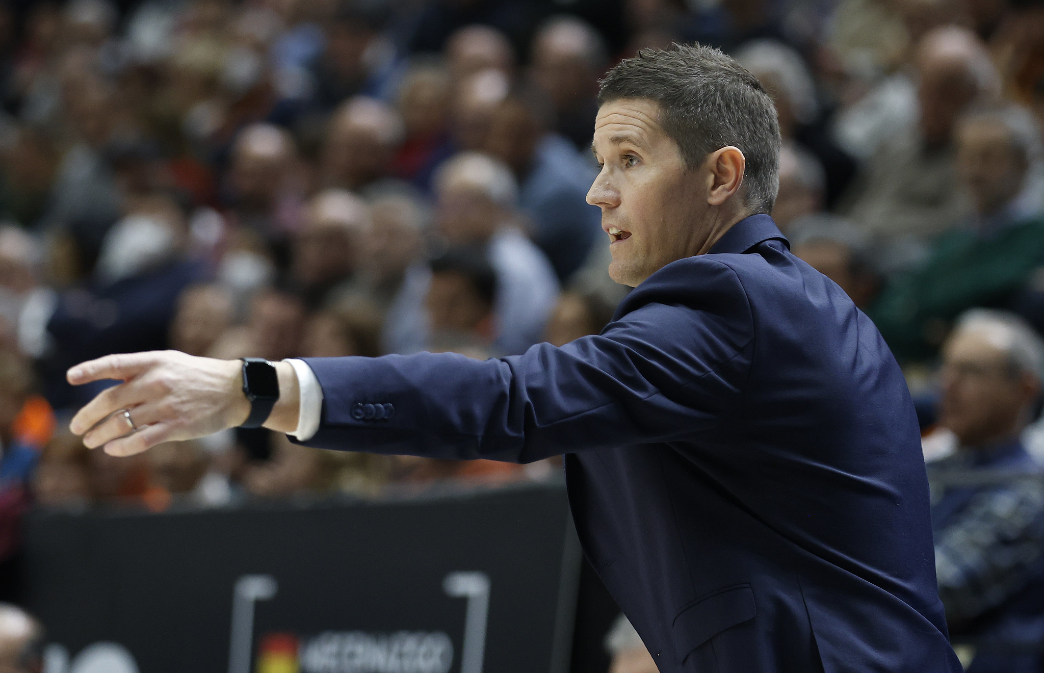 Entrenador dando instrucciones durante un partido de baloncesto, público al fondo.