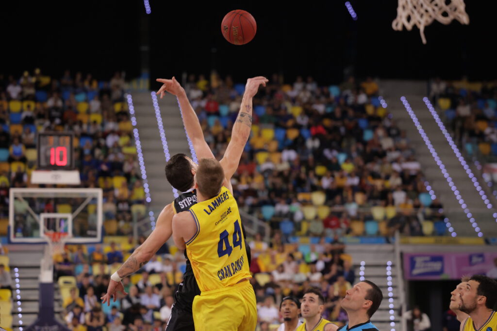 Dos jugadores compiten por el balón en un partido de baloncesto, con el público de fondo.