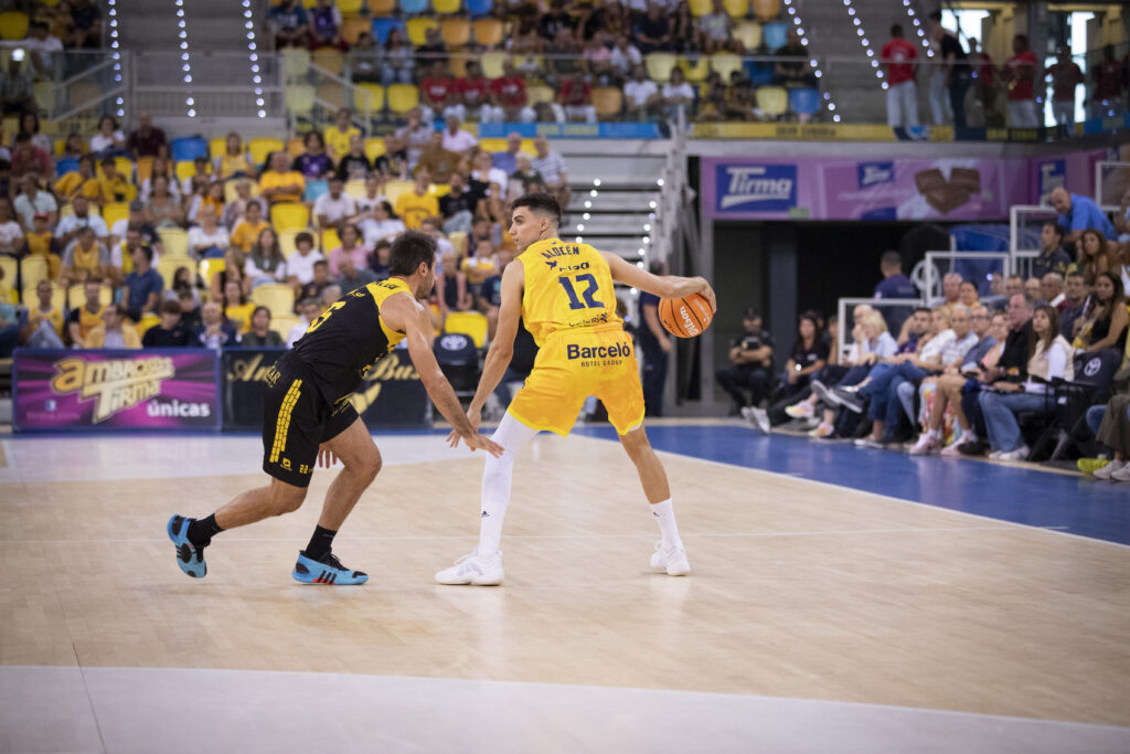 Jugadores en acción durante un partido de baloncesto, uno driblando y el otro defendiendo.