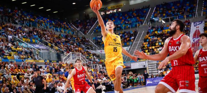 Jugador en acción lanzando el balón hacia la canasta en un estadio lleno de espectadores.
