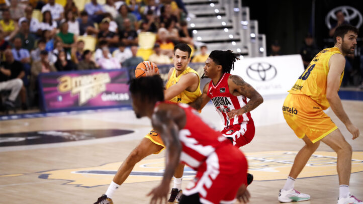 Jugadores de baloncesto en acción durante un partido, con un público animado al fondo.