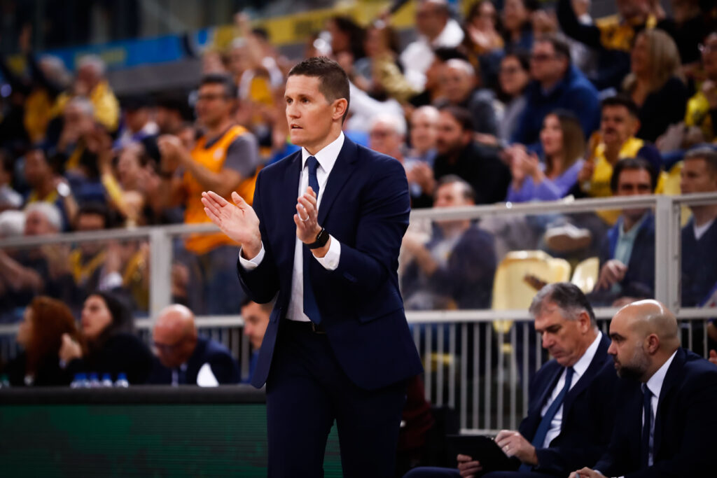 Entrenador aplaudiendo en un partido, con público animando en el fondo.