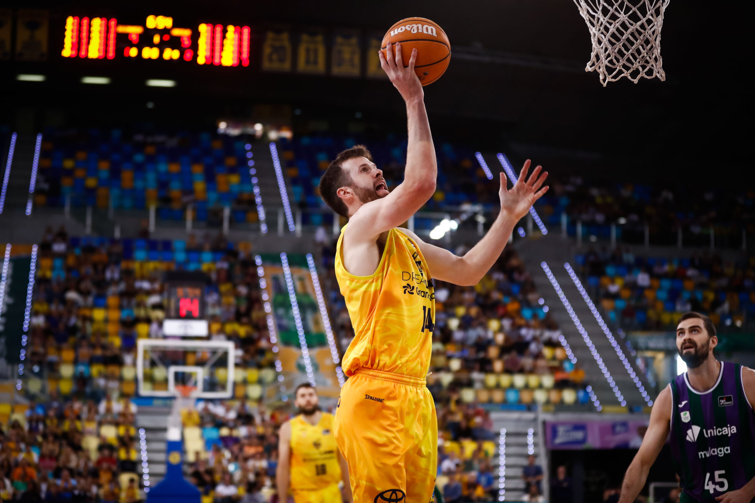 Jugador lanzando a canasta durante un partido de baloncesto. Público en las gradas.