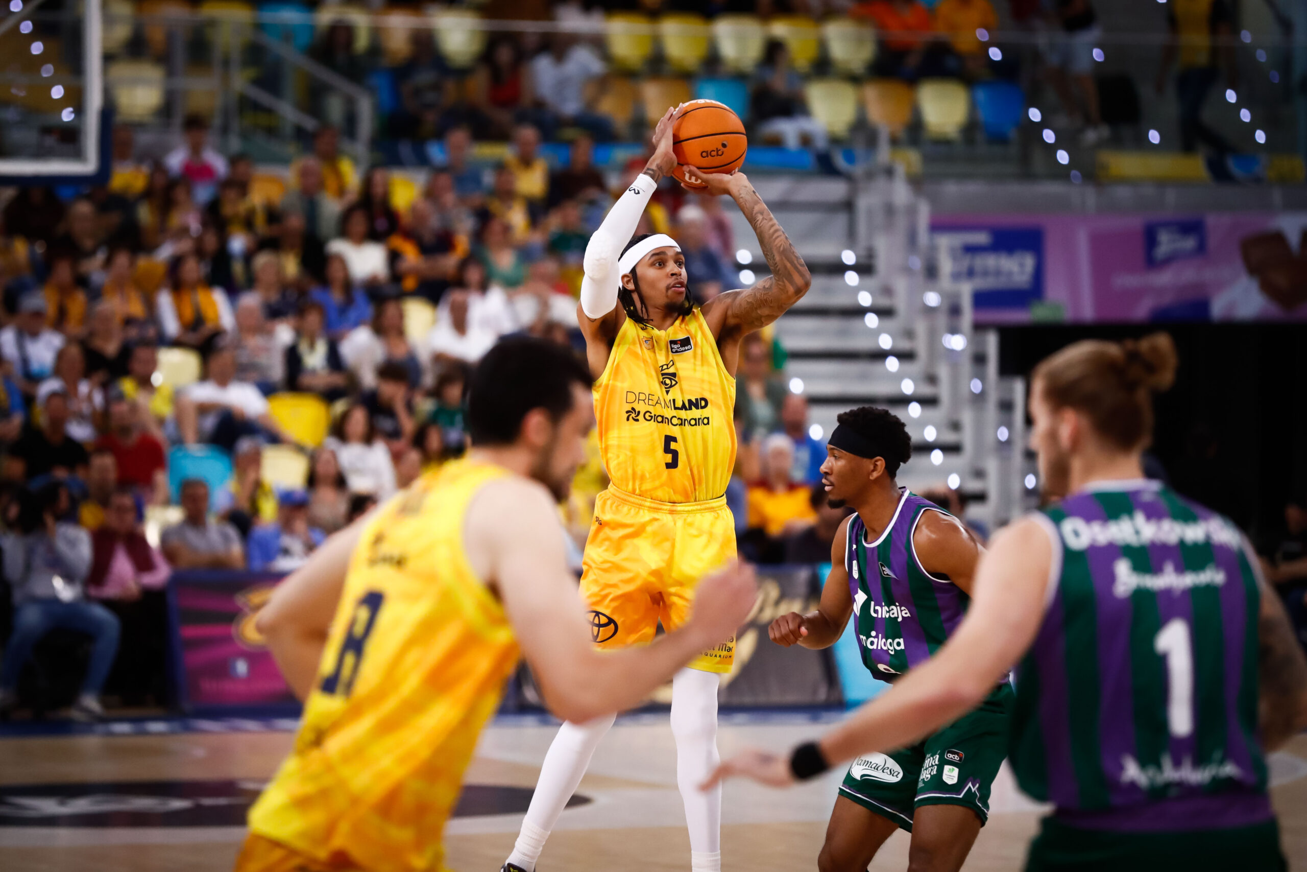 Jugador lanzando a canasta en un partido de baloncesto, con la afición al fondo.