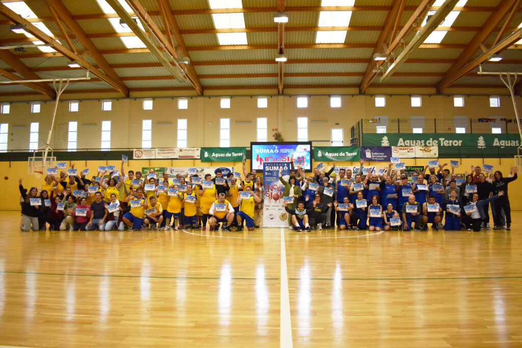 Grupo de personas sonrientes en un evento deportivo, sosteniendo carteles en un polideportivo.