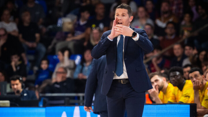 Entrenador dando instrucciones durante un partido, con público animado en el fondo.