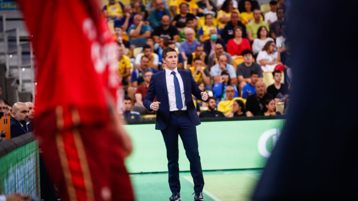 Entrenador en la cancha, gesticulando durante un partido, con aficionados al fondo.