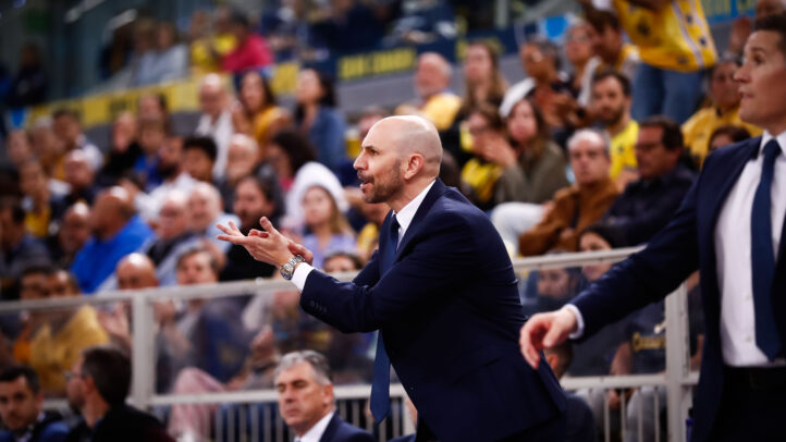 Entrenador dirigiendo al equipo durante un partido, con afición en las gradas.