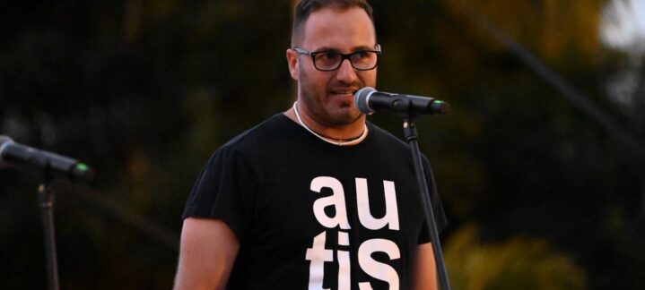 Hombre con camiseta que dice ""autism"" hablando en un micrófono durante un evento.