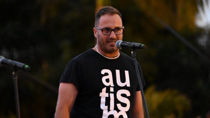 Hombre con camiseta que dice ""autism"" hablando en un micrófono durante un evento.