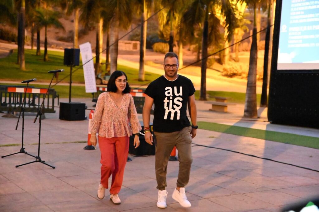 Dos personas caminan por un espacio al aire libre, rodeadas de micrófonos y plantaciones.