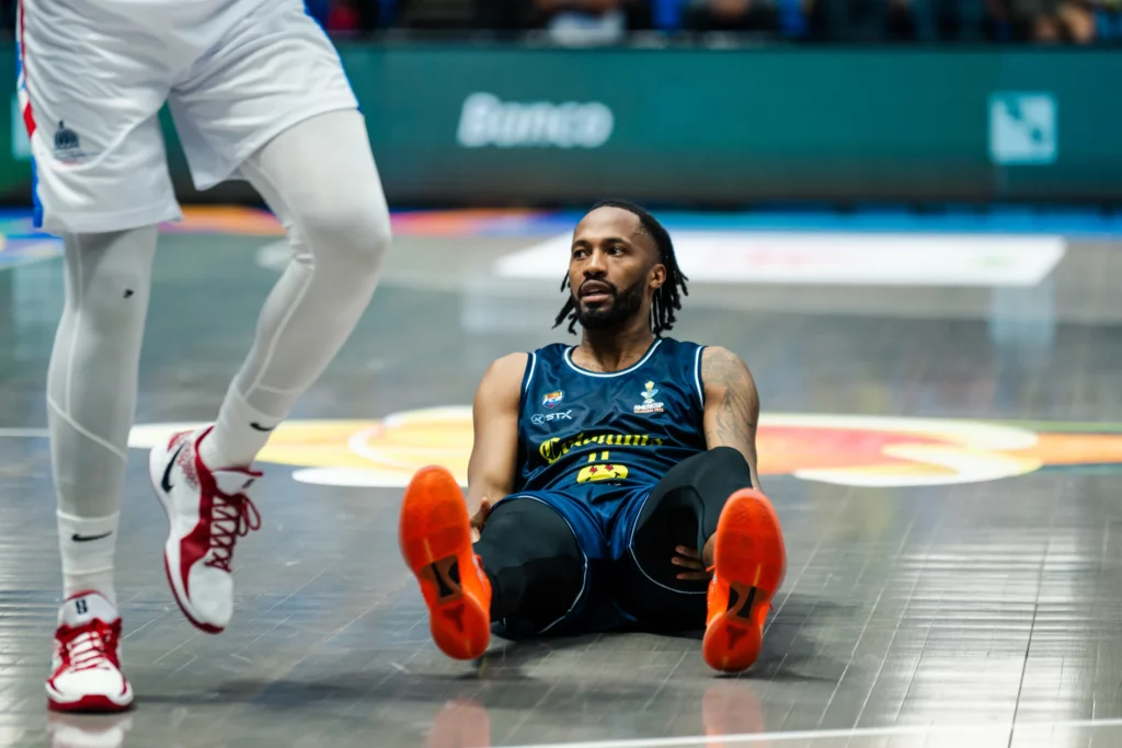 Braian Angola sentado en la cancha, con un jugador de pie a su lado.