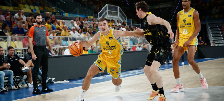 Jugadores en acción durante un partido de baloncesto, con un árbitro e hinchas al fondo.