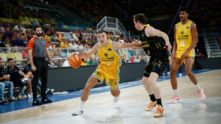 Jugadores en acción durante un partido de baloncesto, con un árbitro e hinchas al fondo.