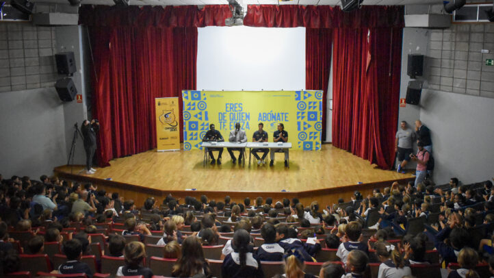 Auditorio con niños aplaudiendo y dos personas en una mesa en el escenario.