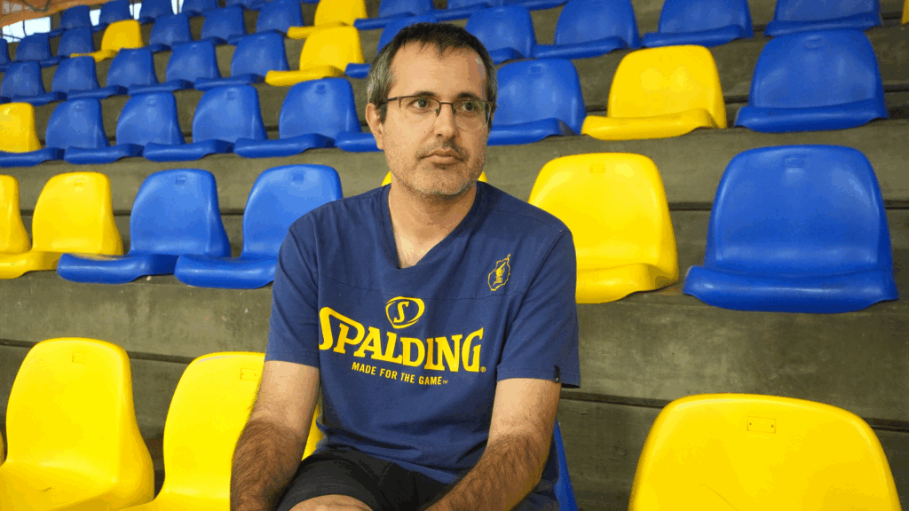 Entrenador sentado en las gradas del pabellón, con asientos amarillos y azules de fondo.