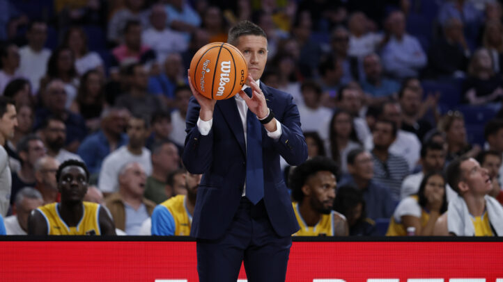 Entrenador sosteniendo un balón de baloncesto y dirigiendo al equipo desde la línea lateral.