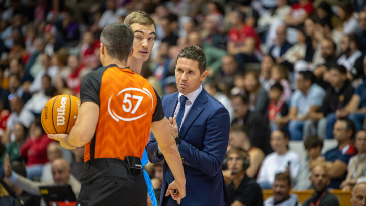 Jaka Lakovic conversa con un árbitro y un jugador durante un partido de baloncesto.