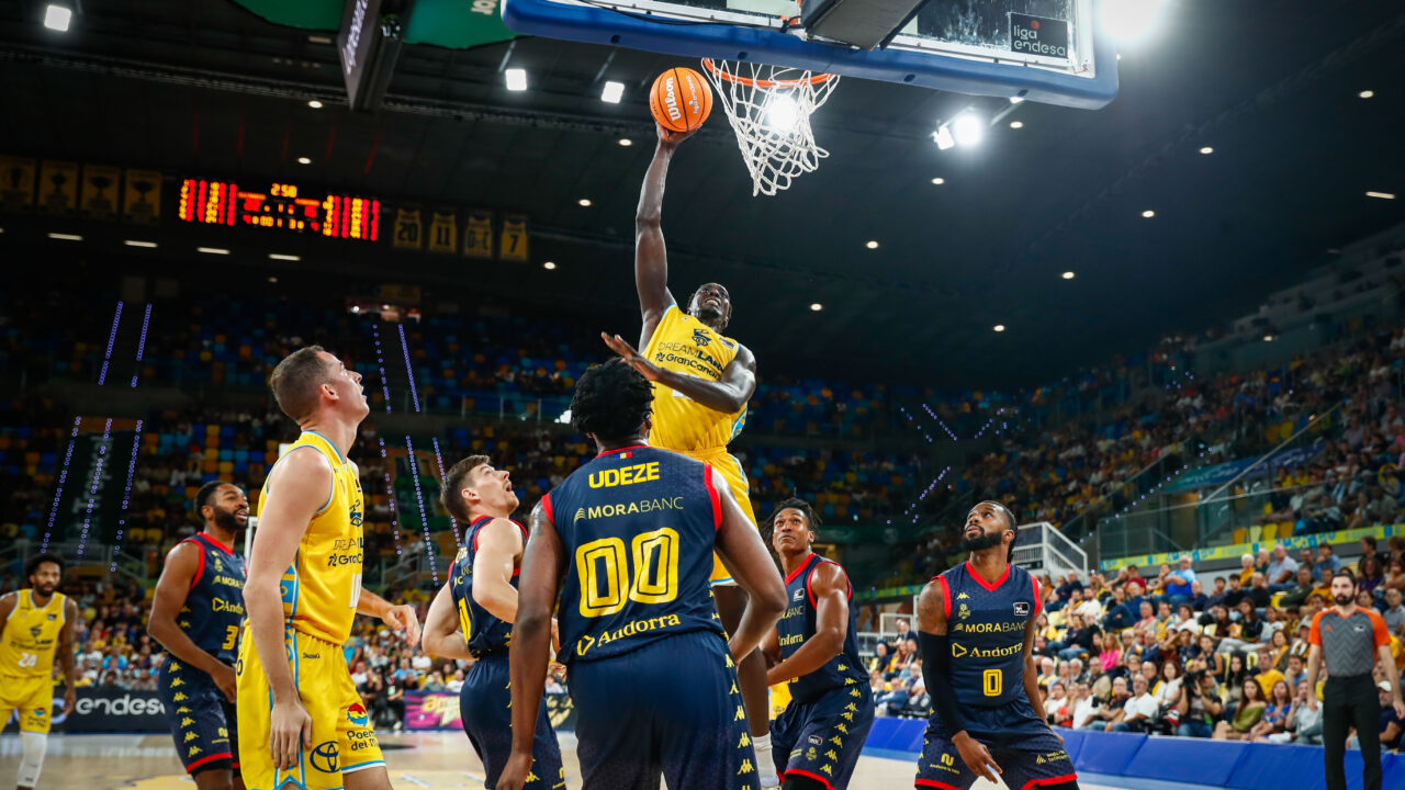 Jugadores de baloncesto compiten en una jugada bajo el aro en un ambiente lleno de público.